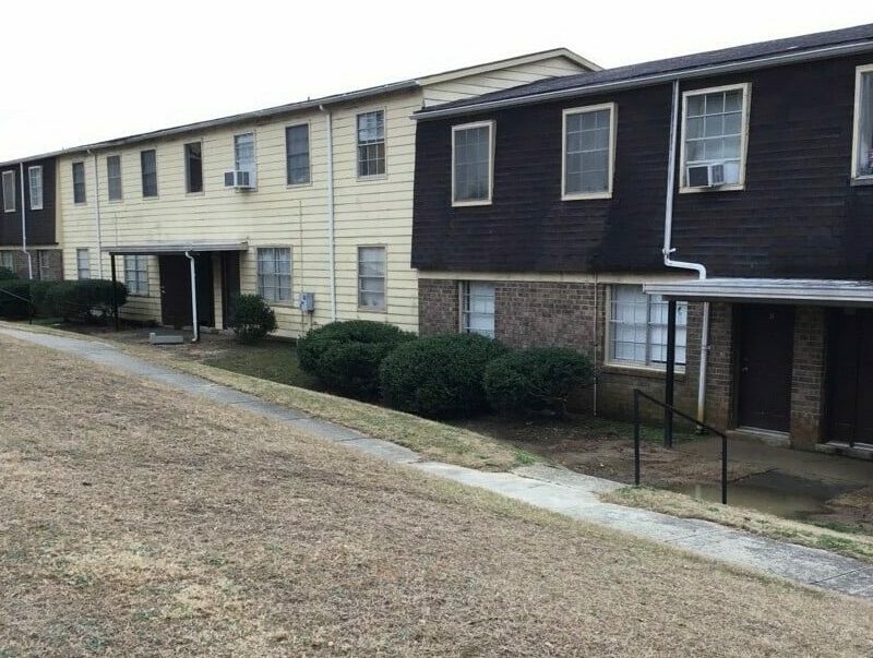 A two-story apartment building with a weathered exterior, featuring light yellow and dark brown siding. The lawn in front of the building is dry and patchy, with a concrete walkway leading to the entrances. Some windows have external air conditioning units, and the shrubs along the building's edge are trimmed low.